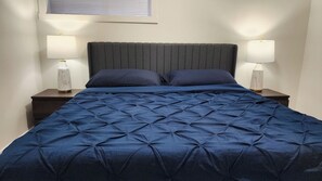 Una cama con colcha azul oscuro, dos lámparas de noche y una ventana con persianas.