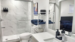 Un baño moderno con cabina de ducha de vidrio, inodoro, lavamanos con espejo y una cama con ropa de cama azul en la habitación contigua.