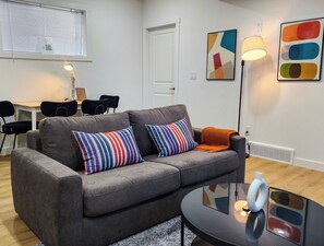 En grå sofa med stribede puder, et lille rundt bord med en hvid genstand, og en væg med abstrakt kunst.