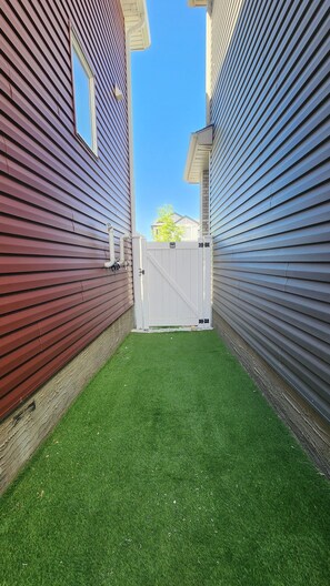 A narrow alleyway with a white gate, green artificial grass, and houses with siding in red and blue. - TNC Stays Cozy Luxurious Modern Suite with 85inch TV (Calgary)