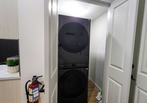 A laundry room with a Samsung washer and dryer, a fire extinguisher, and a potted plant. - TNC Stays Cozy Luxurious Modern Suite with 85inch TV (Calgary)