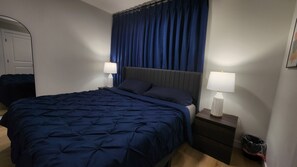 A hotel room with a large bed, two bedside tables with lamps, a dark blue curtain, and a white door. - Modern Suite in Seton - King Bed, 85" TV, Near South Health Campus & YMCA (Calgary)
