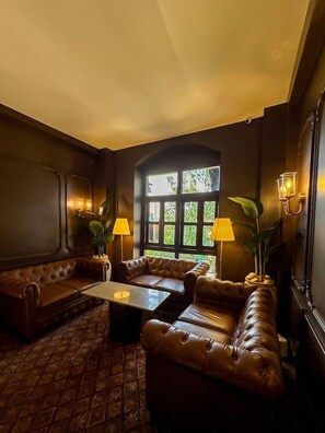 A dimly lit room with a large window, leather sofas, a coffee table, and wall-mounted lamps. - Gulf Hotel (Mumbai)