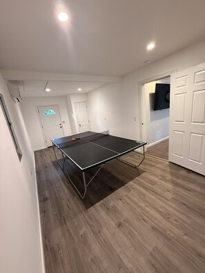 Ein Zimmer mit einem Tischtennis-Tisch, Holzboden, weißen Wänden und einer Tür zu einem weiteren Raum.