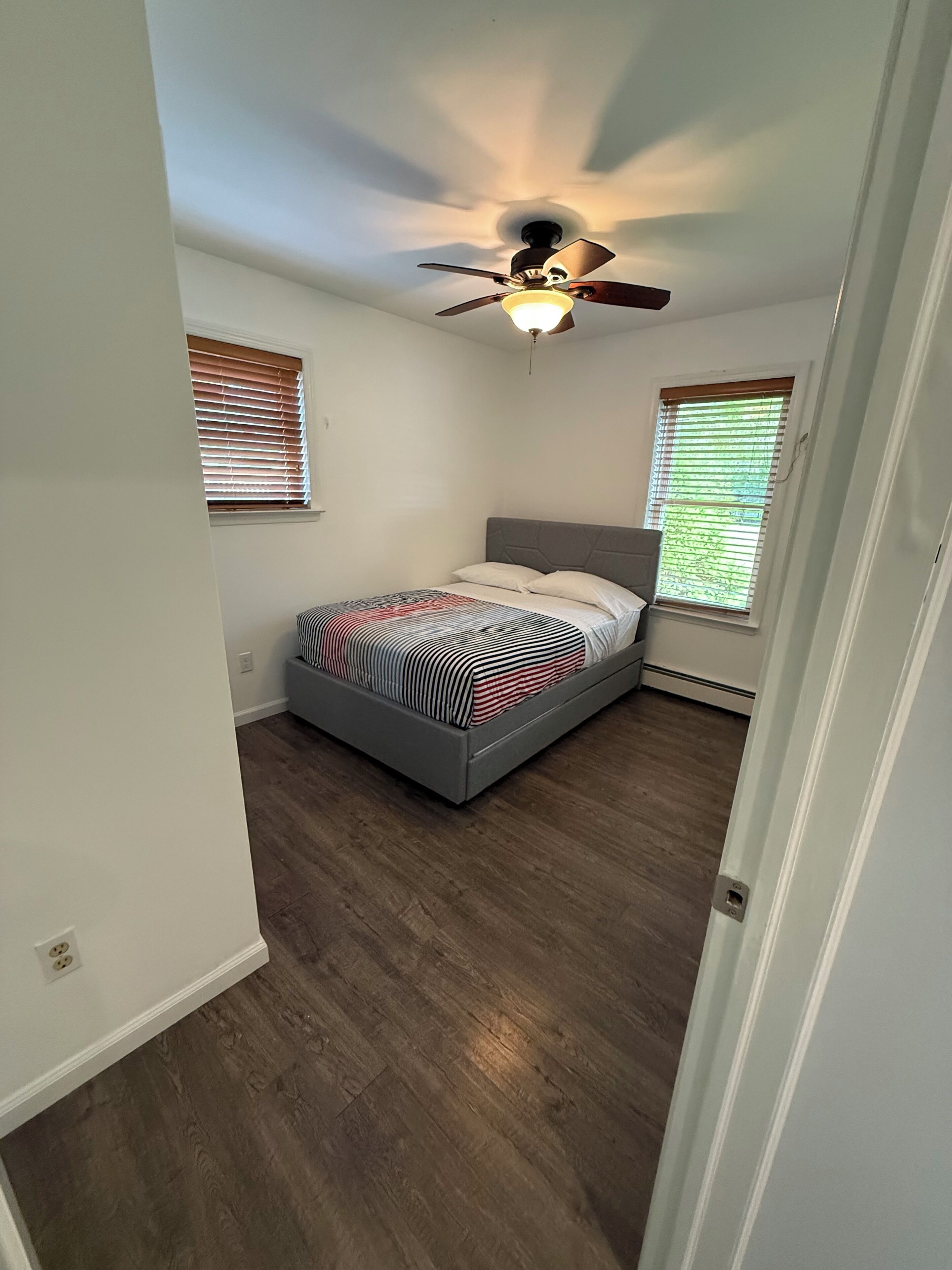 A bedroom with a bed, ceiling fan, and two windows with blinds.