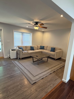 A modern living room with a sectional sofa, a ceiling fan, a coffee table, and a large window with blinds. - 3-bedroom house 5 minutes away from the beach in Hampton Bays (Hampton Bays)