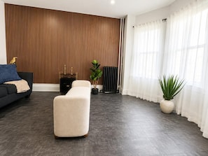 A modern living room with a sectional sofa, a cream-colored ottoman, a potted plant, and a radiator against a wooden panel wall.