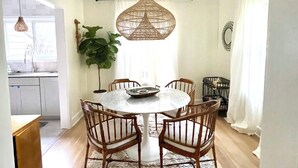 Een eetruimte met een ronde tafel, zes houten stoelen, een geweven hanglamp, een potplant en uitzicht op de keuken.