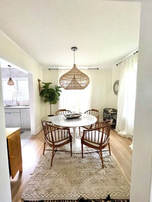 Ein Essbereich mit einem runden Tisch, sechs Holzstühlen, einer gewobenen Pendelleuchte, einem Blumentopf und Blick zur Küche.