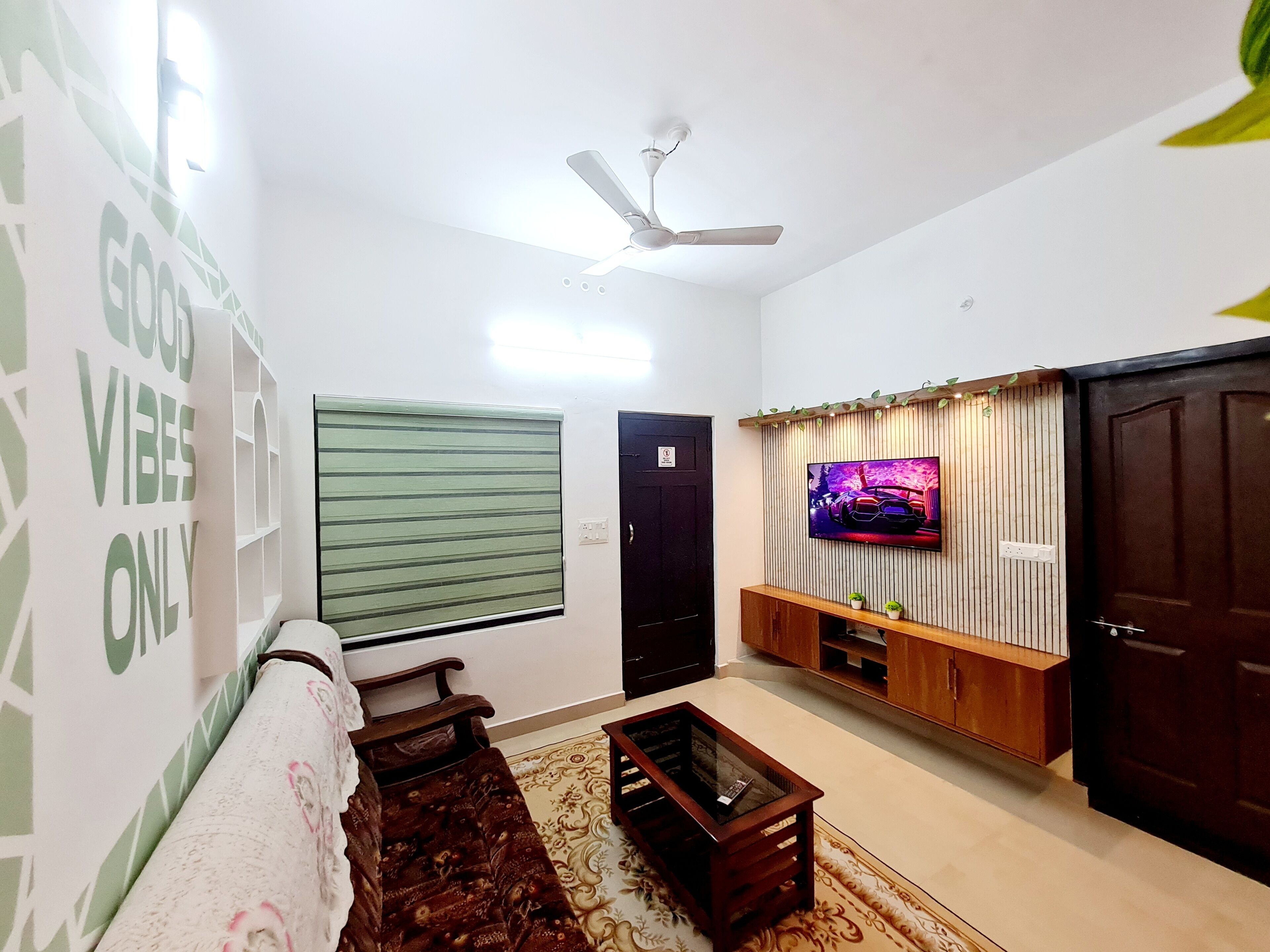 Ein Wohnzimmer mit einer Couch, einem Couchtisch, einem an einer Holzwand montierten Fernseher, einem Deckenventilator und einem Fenster mit Jalousien.