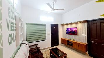 Ein Wohnzimmer mit einer Couch, einem Couchtisch, einem an einer Holzwand montierten Fernseher, einem Deckenventilator und einem Fenster mit Jalousien.
