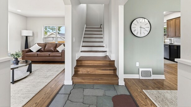 A modern interior with a staircase, a brown leather sofa, a round wall clock, and a kitchen area. - Fully Updated Field Club Colonial - 4BR Near UNMC & Blackstone (Omaha)