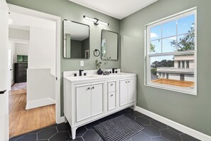 Une salle de bain avec un meuble-lavabo blanc, deux miroirs, un tapis rayé noir et blanc et une fenêtre donnant sur une maison.