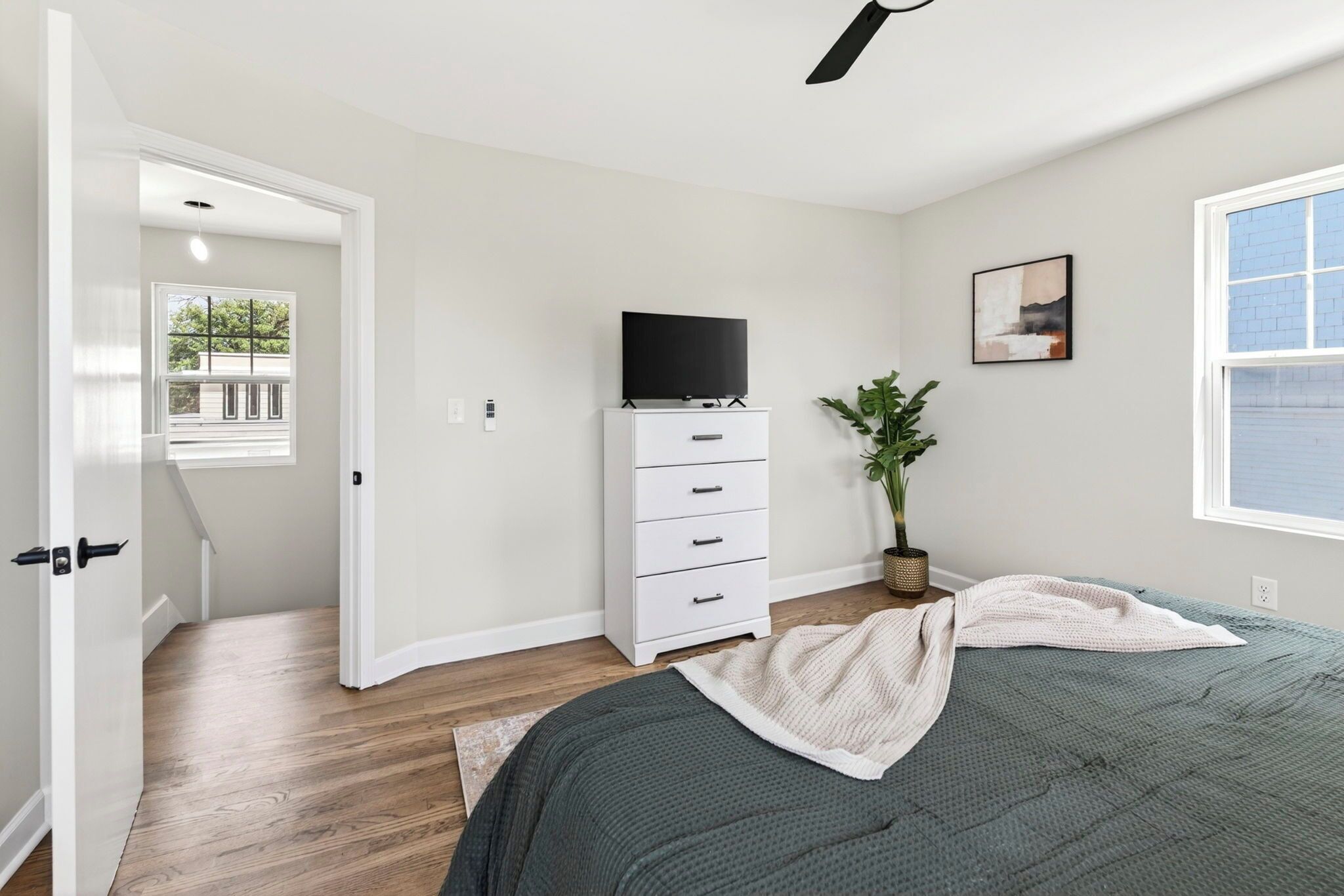 Een slaapkamer met een bed, een dressoir, een televisie erboven, een kamerplant en een raam.