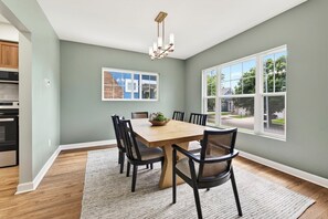 Une salle à manger avec une table en bois, des chaises noires, un lustre, une grande fenêtre et un coin cuisine avec des placards et des appareils électroménagers.