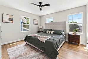 Ein Schlafzimmer mit einem Bett, einem Deckenventilator, zwei Fenstern, einem Nachttisch und einem Teppich.