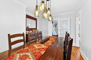 Un comedor con una larga mesa de madera, sillas, lámparas colgantes y un corredor con estampado.