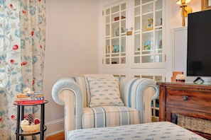 Ein Zimmer mit einem gestreiften Sessel, einem kleinen Tisch mit Büchern, einer Vitrine und einem Fernseher.