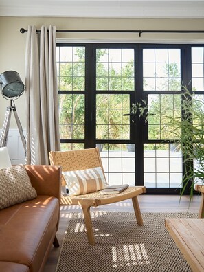 Ein Zimmer mit einem braunen Ledersofa, einem Korbstuhl, einer Stehlampe und einem großen Fenster mit Blick auf Grünflächen.