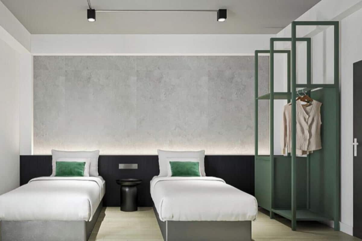 A modern hotel room with two beds, a large wall-mounted light, a small trash bin, and a green wardrobe.