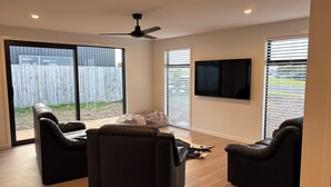 Una sala moderna con un ventilador de techo, un televisor de pantalla plana montado en la pared y un amplio ventanal corredera que da a un jardín.
