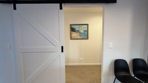 Una habitación con una puerta corrediza de estilo rústico blanca, un cuadro con una playa y dos sillas negras.