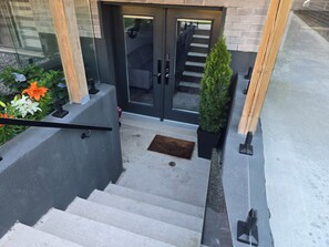 Una escalera conduce a una entrada moderna con puertas de vidrio, una planta pequeña en maceta y un felpudo.