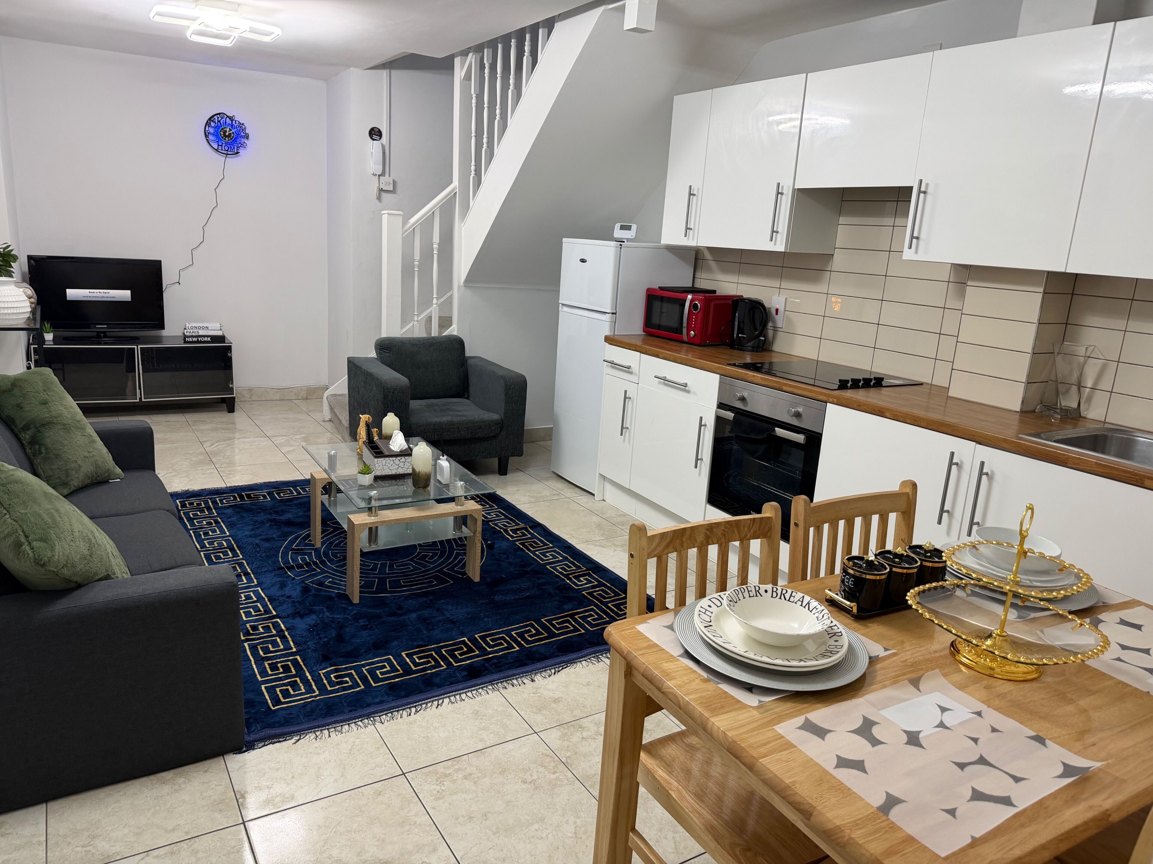 A modern living room with a sofa, coffee table, and a dining table set with plates and a decorative bowl. A kitchen with white cabinets and a microwave is visible.