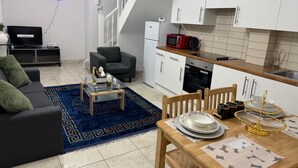 Un salon moderne avec un canapé, une table basse et une table à manger dressée avec des assiettes et un saladier décoratif. On aperçoit une cuisine avec des meubles blancs et un four à micro-ondes.