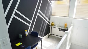 Une pièce avec un mur orné d’un motif géométrique noir et blanc, une petite table avec des chaises, une étagère en verre sur laquelle sont disposés des objets décoratifs, et une fenêtre avec des stores.