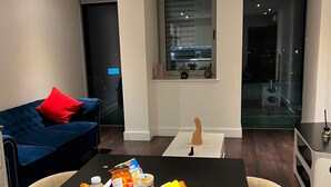 Een moderne woonkamer met een donkerblauwe bank, een klein tafeltje met lekkernijen, een rieten mand en uitzicht op de stad door een groot raam.