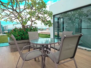 Un patio al aire libre con una mesa de vidrio y cuatro sillas, un árbol con hojas verdes y flores rojas, y una vista de la ciudad a través de un muro de cristal.