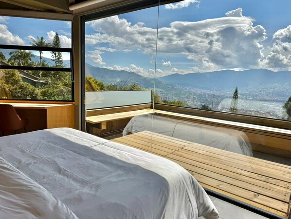 A bedroom with a large window, a bed, a wooden table, and a view of mountains and a city.
