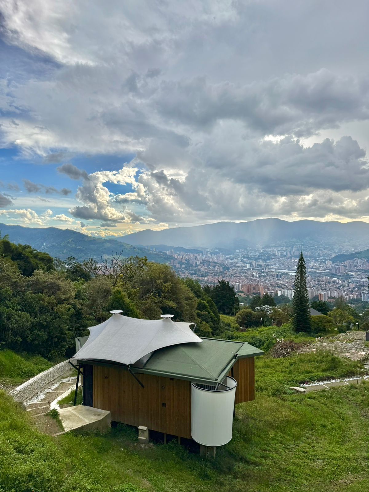 Una cabaña de madera con techo curvo, rodeada de exuberante vegetación y una vista a un paisaje urbano y montañas distantes.