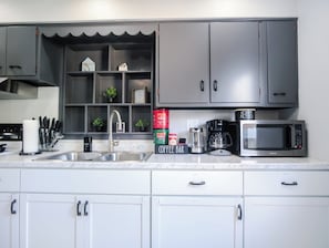 A modern kitchen with grey cabinets, a stainless steel sink, a coffee maker, a microwave, and a coffee bar.