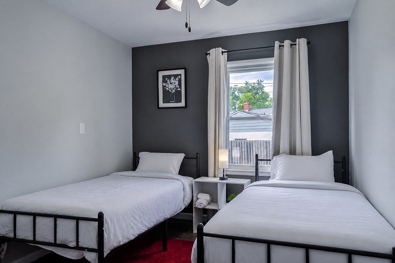 Ein Zimmer mit zwei Einzelbetten, einem kleinen Tisch mit Lampe, einem Fenster mit Vorhängen und einem gerahmten Bild an der Wand.
