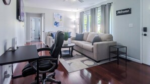 A modern living room with a sofa, coffee table, and a desk with a chair. There is a ceiling fan, a clock, and a 'Welcome' sign on the wall.