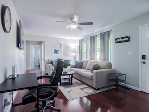 Una sala de estar moderna con un sofá, una mesa de centro y un escritorio con una silla. Hay un ventilador de techo, un reloj y un cartel que dice 'Welcome' en la pared.