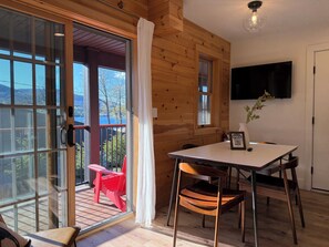 Interior de madera con una puerta corredera de vidrio que da a un balcón, una mesa con sillas y un televisor montado.