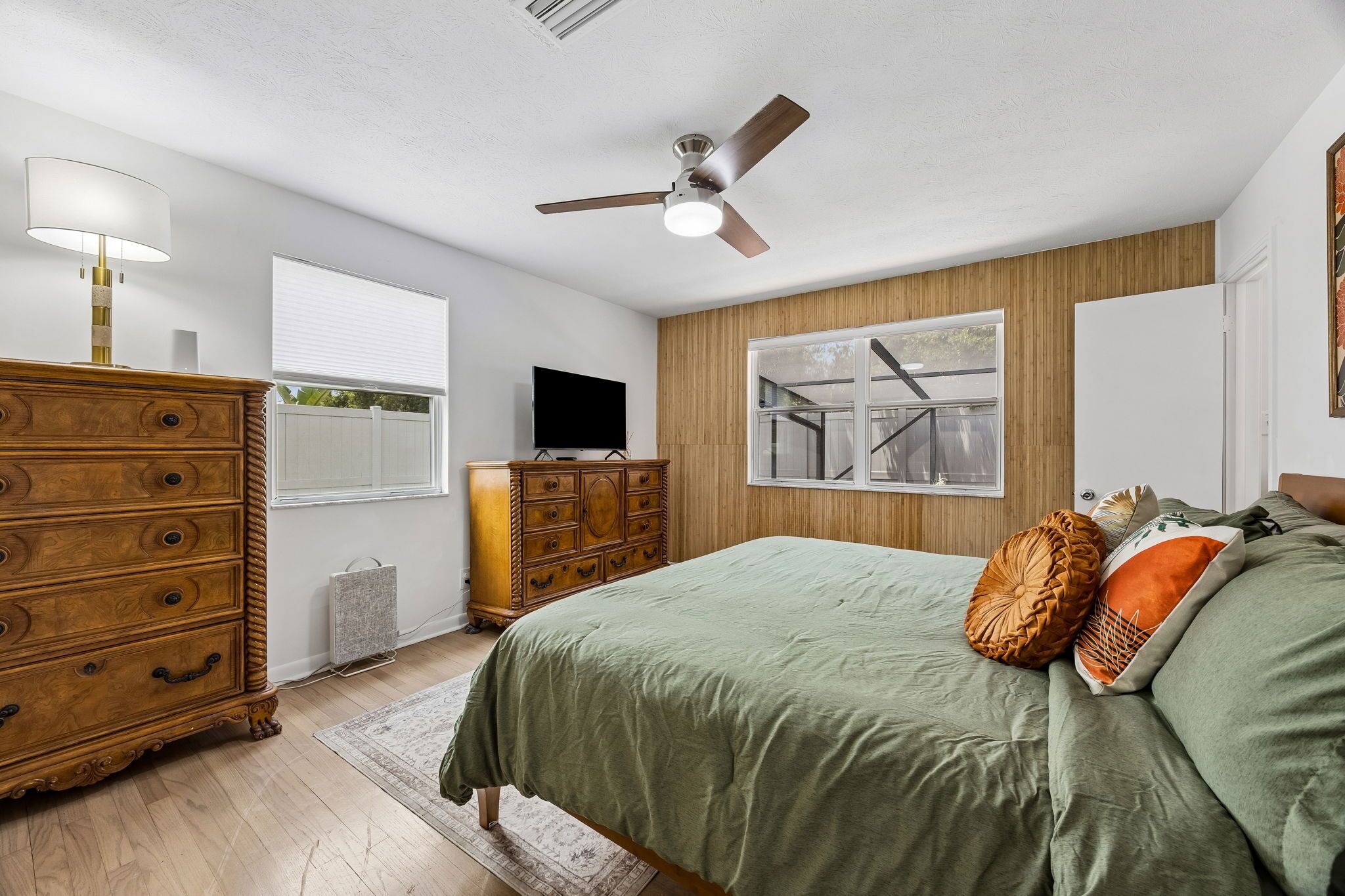 A bedroom with a bed, dresser, TV, ceiling fan, windows, and a view of the outside.