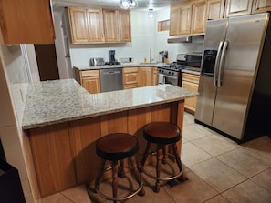 A kitchen with wooden cabinets, a granite countertop, a stainless steel refrigerator, and two stools.