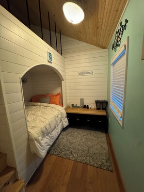 A bedroom with a loft bed, a small desk, a window with blinds, and a wall-mounted sign reading 'BEACH HOUSE'.