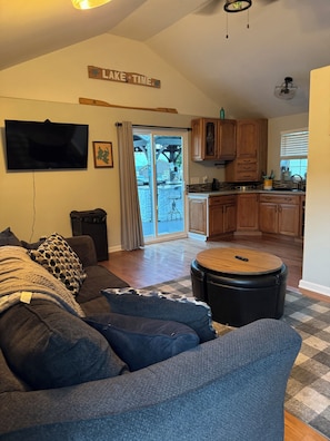 A living room with a gray sectional sofa, a coffee table, a TV, and a kitchen area with wooden cabinets and a stone countertop.