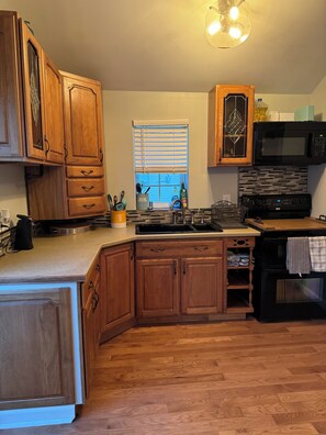 A kitchen with wooden cabinets, a black microwave, a stove, and a window with blinds.
