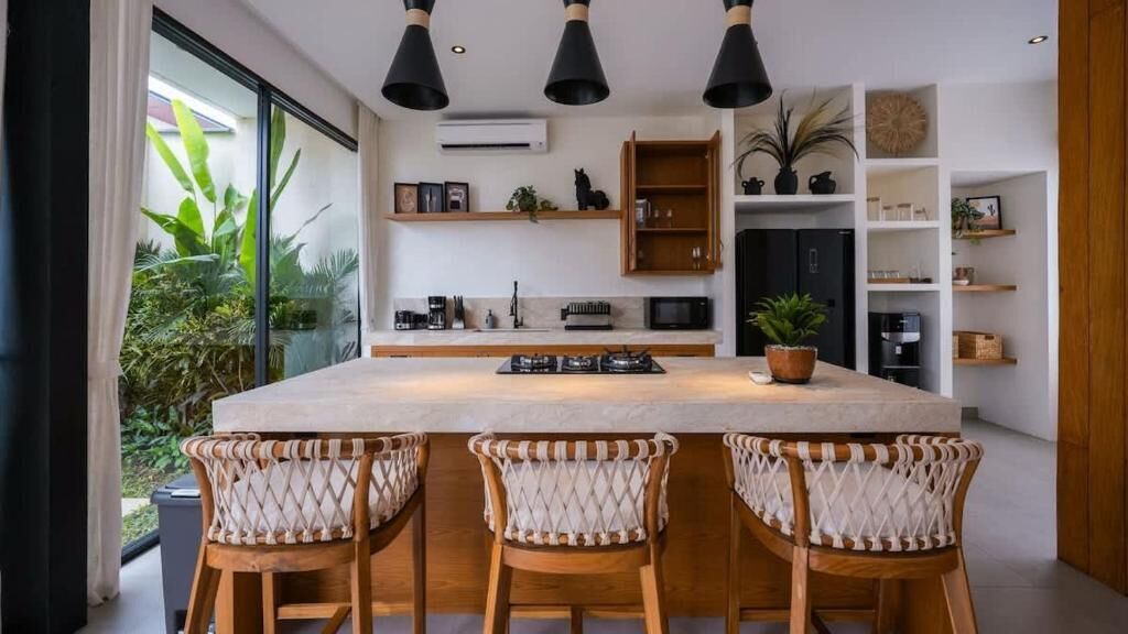 A modern kitchen with a central island, bar stools, and a view of greenery outside.