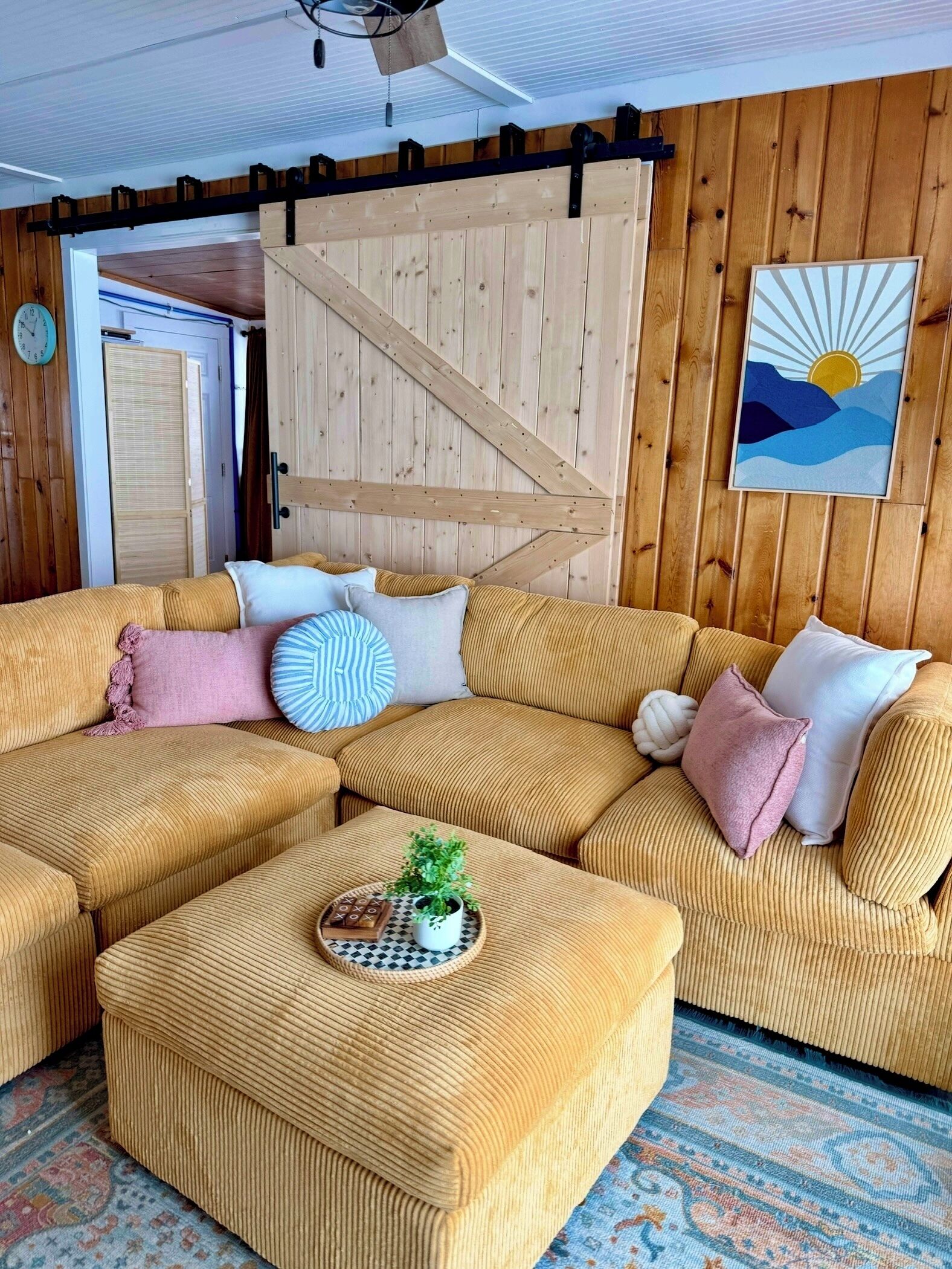 A cozy living room with a large sectional sofa, a small ottoman with a plant and a board game, and a wooden wall with a sun painting.