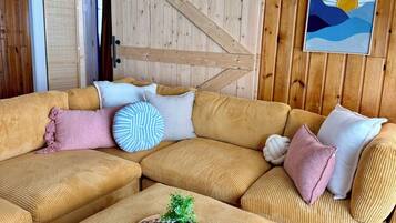 A cozy living room with a large sectional sofa, a small ottoman with a plant and a board game, and a wooden wall with a sun painting.
