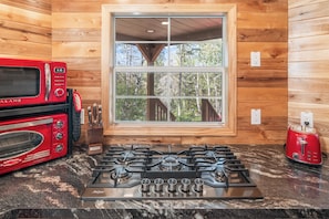 A kitchen with a gas stove, a red toaster, and a window with a view of trees. - Flowing Waters Retreat - Two Cabins with Sauna (Two Harbors)