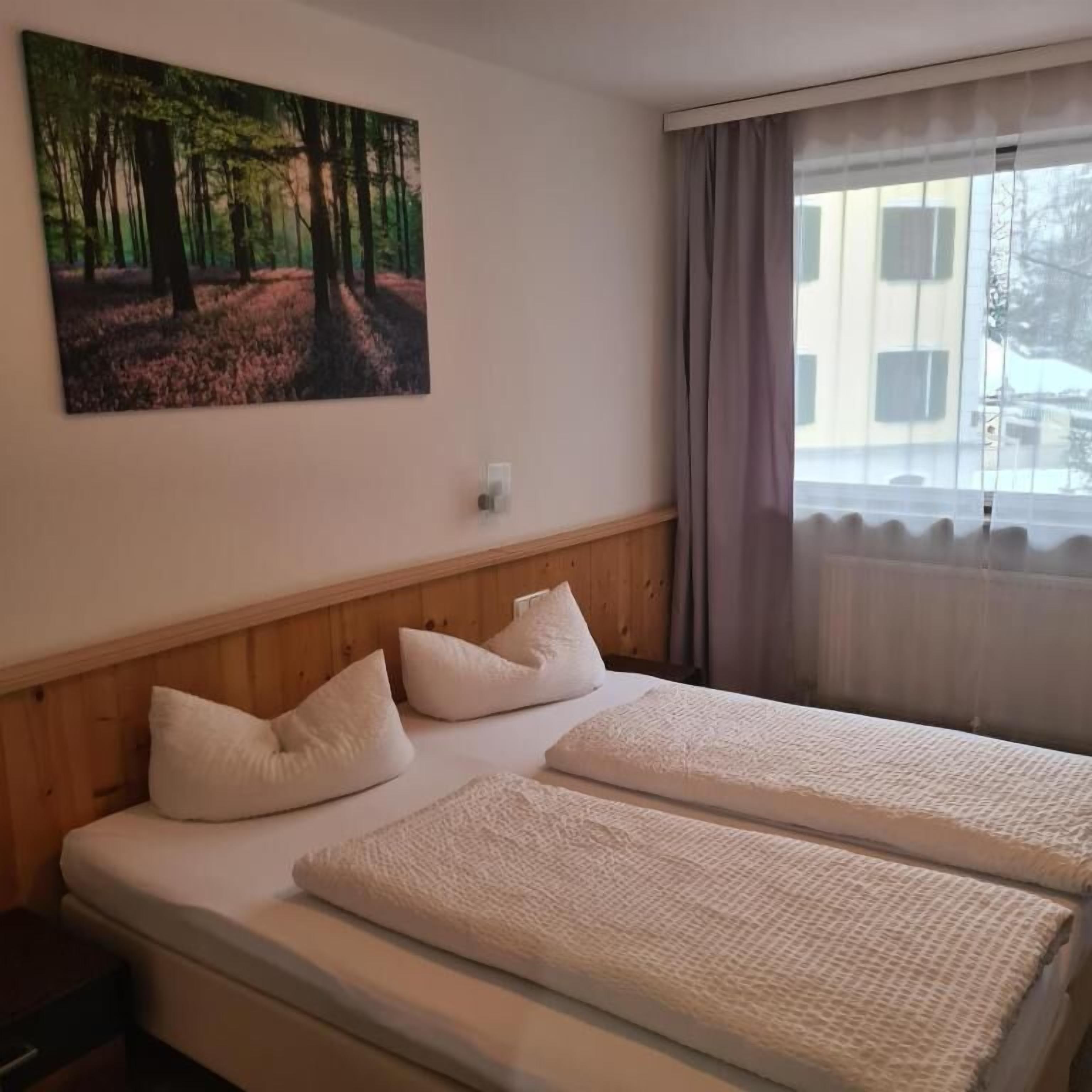 Quarto de hotel com uma cama, um quadro de uma floresta, uma janela com cortinas e vista para edifícios lá fora.