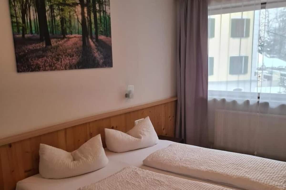 Habitación de hotel con cama, un cuadro de un bosque, una ventana con cortinas y vistas a edificios.